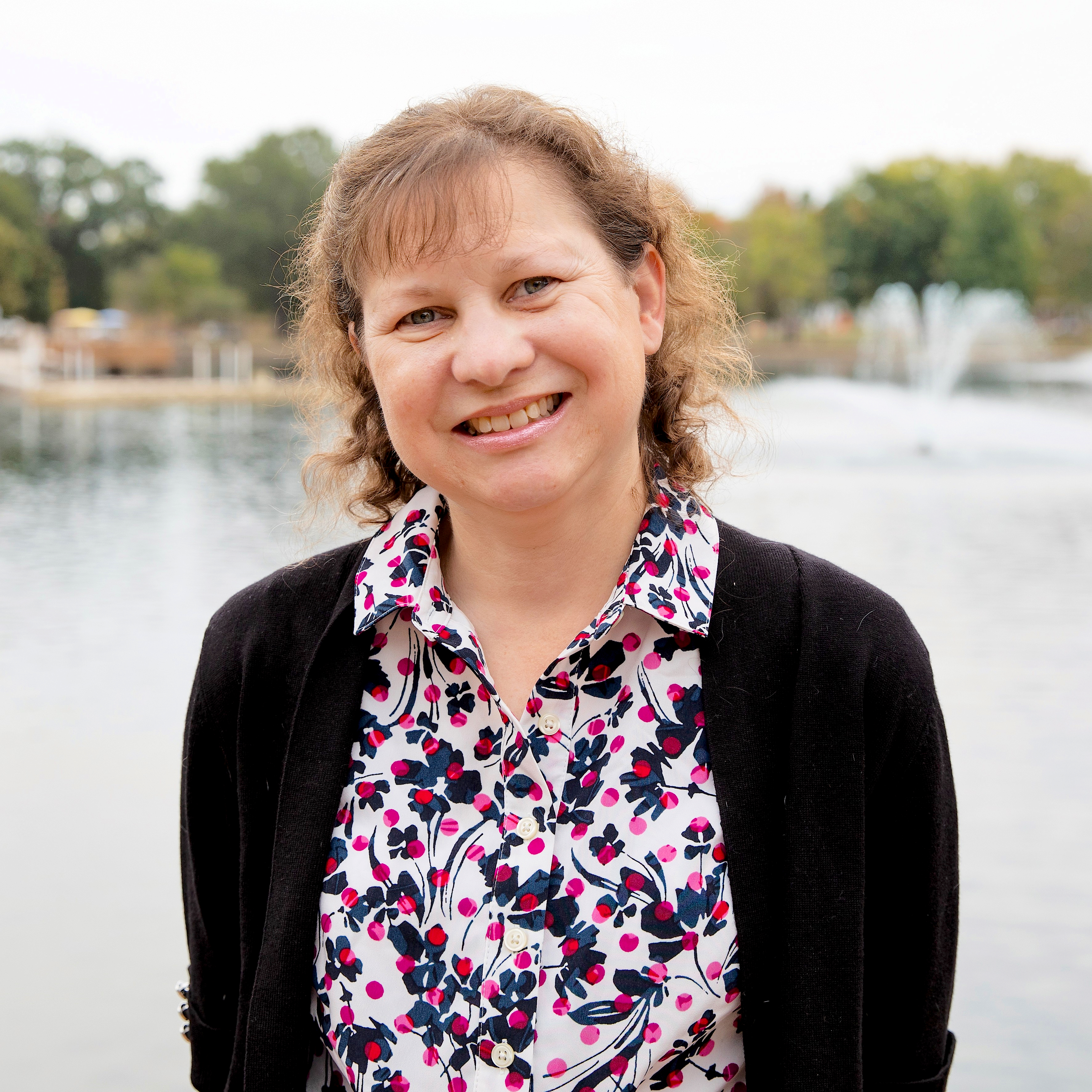 A smiling woman wearing a floral blouse and black cardigan is standing in front of a body of water.