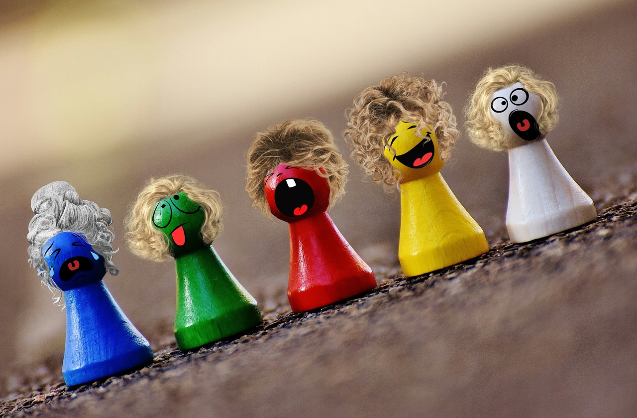 Six wooden game pieces are lined up side-by-side. Each piece has a round, tapered base with a sphere at the top. Each piece is a different color and has a different emoji expression on the sphere to create a "face" for the game mover. All the movers have wigs, making them look more like people.