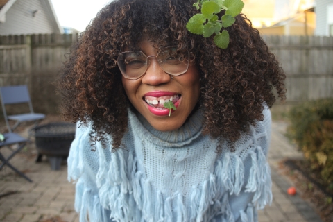 A smiling black woman wearing glasses has a sprig of leaves in her hair and a single leaf hanging out of her mouth.