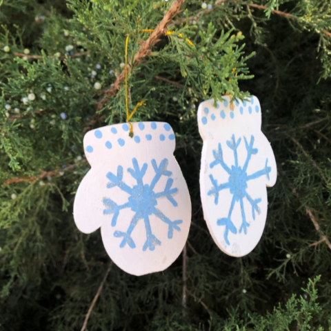 Wooden ornaments in the shape of mittens painted with a blue snowflake and dots on a white background hanging in a tree.